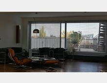 Modernes Wohnzimmer mit minimalistischer Einrichtung, bestehend aus schwarzen Ledersesseln, einem Holz-Lounge-Stuhl mit Hocker und einem Couchtisch. Im Hintergrund große Fenster mit Jalousien, die auf eine Terrasse mit Pflanzen und einer Wendeltreppe führen.