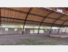 Offene Halle mit einem gewölbten Dach aus hellem Holz, das von schwarzen Metallträgern gestützt wird. Die Seitenwände bestehen aus teilrenovierten Ziegelmauern mit Fensteröffnungen; der Boden ist mit Erde und vereinzeltem Gras bewachsen.