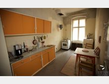 Einfache Küche mit orangefarbenen Schränken, einem weißen Herd und einem kleinen Tisch mit zwei Stühlen. Die Wände sind teils unverputzt, ein großes Fenster sorgt für Tageslicht, und ein kleiner Kühlschrank steht in der Ecke.