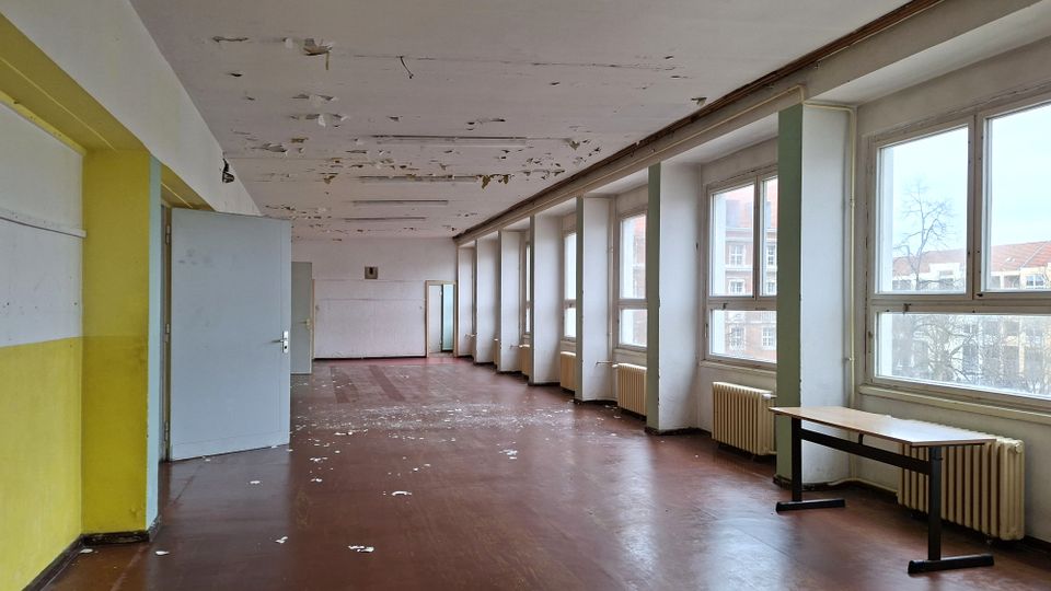 Langer, leerer Innenraum eines älteren Gebäudes mit großen Fensterreihen entlang der rechten Seite, Heizkörpern darunter und braunem Bodenbelag. Die Decke zeigt abblätternde Farbe, und am Ende des Raums stehen offene Türen sowie ein einzelner Tisch nahe den Fenstern.