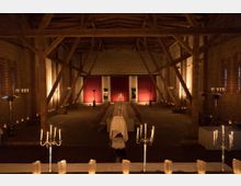 Eine langgestreckte Veranstaltungshalle mit Sichtmauerwerk aus Ziegelsteinen, sichtbaren Holzbalken und einem hölzernen Dachstuhl. Im Raum steht eine lange, gedeckte Tafel mit Stühlen, im Hintergrund hängen rote Vorhänge, und der Raum ist mit Kerzenleuchtern und indirektem Licht stimmungsvoll beleuchtet.