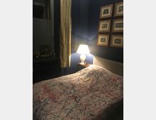 Gemütliches Schlafzimmer mit dunkelblauen Wänden, einem Bett mit farbgesprenkelter Bettdecke und einem Nachttisch mit leuchtender Tischlampe. Links ein Fenster mit Vorhang und Fensterbank, rechts eine Wand mit mehreren gerahmten Bildern.
