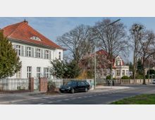 Straßenszene in einem Wohngebiet mit freistehenden Häusern in weißem Putz und roten Ziegeldächern, umgeben von Vorgärten und Bäumen. Im Vordergrund verläuft ein Gehweg mit einem parkenden Auto, sowie einer Straße mit Markierungen und einer Straßenlaterne.