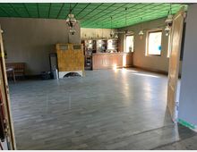 Ein leerer Raum mit grauem Laminatboden, grün gestrichener Decke und mehreren aufgehängten Lampen. Im Hintergrund befindet sich eine Holztheke mit Barregal, daneben ein gelber Kachelofen sowie mehrere Fenster, die Tageslicht hereinlassen.