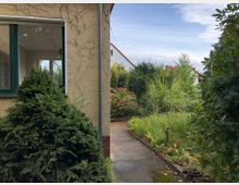 Eingangsbereich eines Gartens mit einem Weg, der zwischen dichter Vegetation verläuft, darunter Nadelsträucher, Blütenpflanzen und hohes Gras. Links ist eine Hauswand mit beige Putz und einem Fenster mit grünem Rahmen zu sehen.