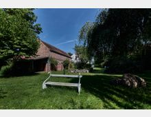 Backsteinbau mit Satteldach in ländlicher Umgebung, umgeben von Bäumen und Rasenflächen. Im Vordergrund steht eine weiße Holzbank, und im Hintergrund sind Sträucher und ein lockerer Steingarten zu sehen.