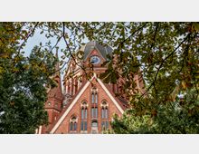 Eine gotische Backsteinkirche mit spitz zulaufenden Türmen und einer Uhr auf dem Hauptturm, teils verdeckt von grünen Baumzweigen im Vordergrund.