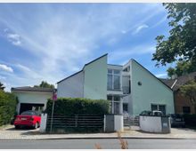 Modernes Wohnhaus mit heller Fassade und asymmetrischem Giebeldach. Ein rotes Auto steht in einer Einfahrt, umgeben von einer niedrigen Hecke und Metallzaun, mit einem „Halteverbot“-Schild entlang der gepflasterten Straße im Vordergrund.