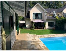 Ein gepflegter Garten mit einer cremefarbenen modernen Doppelhaushälfte im Hintergrund, ausgestattet mit einer Terrasse, großen Fenstern und einem kleinen Balkon. Im Vordergrund befindet sich ein Pool mit hellen Steinplatten und eine grüne Rasenfläche, umgeben von Gartenmöbeln und Pflanzen.