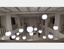 Blick in ein zweigeschossiges, helles Atrium mit umlaufenden Galerien und balustradenartigen Brüstungen an den Seiten. Von der hohen Decke hängen zahlreiche runde Kugelleuchten in unterschiedlichen Größen an langen Kabeln in den offenen Raum.