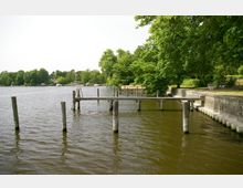 Holzsteg am Ufer eines Sees, umgeben von üppigen, grünen Bäumen. Im Hintergrund sind weitere Stege, Boote und Gebäude sichtbar, während eine gemauerte Uferbefestigung rechts entlangläuft.