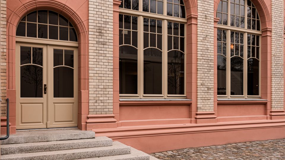 Historische Gebäudefassade in Rosé mit großen Rundbogenfenstern und dekorativen Stuckelementen zwischen hellen Ziegelpfeilern. Links führt eine kurze Steintreppe zu einer zweiflügeligen Eingangstür, davor liegt ein gepflasterter Innenhof aus Kopfsteinpflaster.