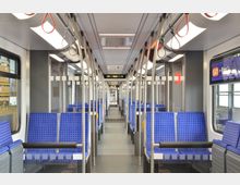 Innenraum einer modernen Stadtbahn mit blauen, gepunkteten Sitzpolstern, Stehhaltestangen aus Metall, weißen Beleuchtungselementen an der Decke und elektronischen Anzeigetafeln. Seitlich befinden sich große Fenster, und oberhalb der Sitze hängen rote Halteschlaufen.