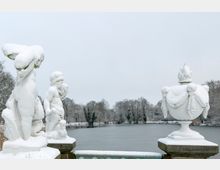 Schneebedeckte Steinfiguren und eine verzierte Vase stehen auf einem Geländer mit Blick auf einen zugefrorenen See, umgeben von kahlen, winterlichen Bäumen. Die Szene ist von einer ruhigen, verschneiten Parklandschaft geprägt.