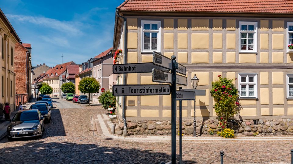 Kopfsteingepflasterte Altstadtstraße mit parkenden Autos und historischen Gebäuden, darunter ein zweigeschossiges Fachwerkhaus mit roten Ziegeldach und Blumenkästen. Im Vordergrund steht ein Wegweiser mit Hinweisen unter anderem zum Bahnhof und zur Touristinformation.