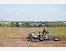Eine verbrannte Parkbank steht auf einer offenen, teils grasbewachsenen Fläche mit Blick auf eine Großstadt im Hintergrund. Umgeben von Bäumen sind in der Ferne Wohnhäuser und ein Gebäude mit großflächiger Werbung sichtbar.