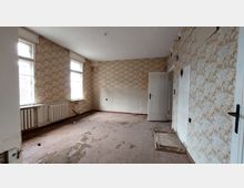 Verlassener Raum mit bröckelndem Holzboden, vergilbter Tapete mit floralem Muster und zwei großen Fenstern, die schwaches Tageslicht hereinlassen. Ein weiß gestrichener Heizkörper unter einem der Fenster, sowie eine geöffnete Tür und sichtbare Schäden an den Wänden und Boden betonen den heruntergekommenen Zustand.