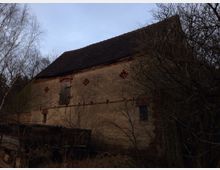 Verfallenes Gebäude aus Backstein mit dunklem Ziegeldach, teilweise bewachsen von kahlen Bäumen. Die Fassade zeigt kleine Zieröffnungen in Backsteinmustern, eine verschlossene Holzluke und ein vergittertes Fenster.