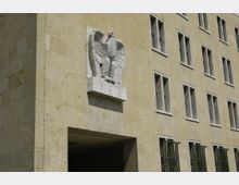 Fassade eines historischen Gebäudes aus heller Steinverkleidung mit regelmäßigen rechteckigen Fenstern. Über einem Durchgang ist ein Relief eines stilisierten Adlers angebracht.