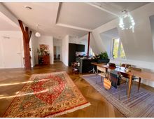 Großzügiger Wohnraum mit offenem Grundriss, schrägen Wänden und großen Fenstern, die viel Tageslicht hereinlassen. Der Raum ist mit einem Holzboden, einem roten Orientteppich, einem Esstisch mit Stühlen und Holzbalken als sichtbare Konstruktionselemente ausgestattet.