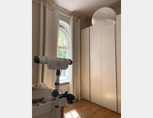 Ein heller Raum mit einem Holzfußboden, einem großen weißen Kleiderschrank und einem runden, weißen Deckenlicht. Im Vordergrund steht ein Babywickeltisch, daneben ein Fenster mit weißen Vorhängen, durch das Tageslicht einfällt.