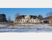 Herrenhaus im Barockstil mit cremefarbener Fassade und schwarzem Mansarddach, umgeben von schneebedecktem Gelände und Bäumen; im Vordergrund ein eingezäuntes Grundstück mit einem Wohnwagen.