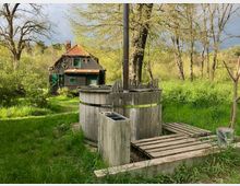 Holzbadefass mit einem Schornstein auf einer Wiese, umgeben von grüner Vegetation und Bäumen; im Hintergrund ein altes, verwittertes Haus mit rotem Ziegeldach und grünen Fensterläden.