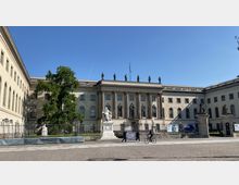 Historisches Gebäude der Humboldt-Universität in Berlin mit klassizistischer Fassade, großen Fenstern und Säulen. Eine Statue steht im Vordergrund auf einem Sockel, während eine große grüne Baumkrone den linken Bereich des Bildes dominiert.