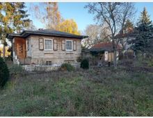 Einfamilienhaus mit Holzfassade und Fensterläden, umgeben von einem verwilderten Garten mit Bäumen und Sträuchern; im Hintergrund weitere Gebäude mit roten Ziegeldächern. Die Szene befindet sich in einer ruhigen, ländlichen Umgebung.