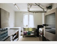 Gewerbliche Küche mit gefliesten Wänden und Böden, ausgestattet mit Edelstahlarbeitsflächen, Großgeräten wie einem Hobart-Geschirrspüler, einem Backofen und einer mobilen Arbeitsstation mit Körben unter einem großen Fenster. Eine Schienenkonstruktion hängt an der Decke.