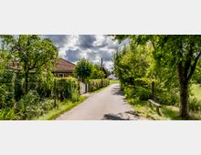 Ländlicher Weg mit Bäumen und Sträuchern an den Seiten, flankiert von einem Backsteinhaus mit rotem Ziegeldach auf der linken Seite. Im Hintergrund sind weitere Häuser und Wiesen zu sehen, unter einem teils bewölkten Himmel.