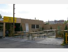 Ein einfacher Außenbereich einer Imbissbude mit Holzbänken und -tischen auf einem Betonboden. Die Imbissbude besteht aus einer Holzkonstruktion mit Wänden aus Schilfrohr, einem gelben Schild mit Speisenangebot und mehreren Pflanzen in Holzkübeln als Dekoration.