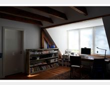 Ein gemütliches Arbeitszimmer unter einer schrägen Decke mit freiliegenden Holzbalken. Links steht ein Regal mit Büchern und Aufbewahrungsboxen, rechts ein Schreibtisch mit Computer und Zubehör vor einem großen Fenster mit Jalousien, das Tageslicht hereinlässt.