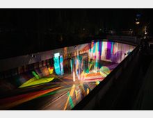 Kunstinstallation auf einem Schiff mit mehreren transparenten, geometrischen Elementen, die mit buntem Licht angestrahlt werden und leuchtende Farbreflexionen auf den Wänden und Holzboden erzeugen. Im Hintergrund sind Bäume und schwach beleuchtete Gebäude zu erkennen.