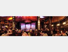 Ein luxuriöser Veranstaltungsraum mit runden Tischen, weißen Tischdecken und Kerzenbeleuchtung, darunter stehen Stühle. Im Hintergrund befindet sich eine beleuchtete Bühne mit glitzerndem Vorhang, umgeben von Holzsäulen und dekorativen Spiegelwänden.