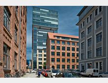 Straßenszene mit alten Backsteingebäuden und modernen Glasbauten, umgeben von geparkten Autos und Fahrrädern. Im Hintergrund ragt ein hochmodernes Gebäude mit Glasfassade empor, während im Vordergrund Backstein- und Putzfassaden mit großen Fenstern zu sehen sind.