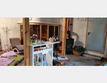 Gemütlicher Innenraum mit einer kleinen Sitzecke, einem Holzofen, einer Sauna mit Glasfront und dekorativen Elementen wie Blumenvasen und einer Buddha-Statue. Der Boden besteht aus Pflastersteinen, und eine Glastür führt in einen weiteren Raum.