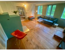 Gemütliches Wohnzimmer mit breiten Holzdielen, weiß verputzten Wänden und großen, grün gestrichenen Sprossenfenstern mit Blick ins Grüne. Ein schwarzes Ledersofa mit kleinem Couchtisch, eine Ottomane sowie ein gemauerter grüner Ofen und ein Durchgang in einen Nebenraum prägen den Raum.