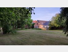 Weitläufiger Garten mit Rasenfläche, umgeben von Bäumen, im Hintergrund ein Backsteinhaus mit grauem Satteldach und angrenzende Gebäude. Der Himmel ist klar und leicht bläulich.