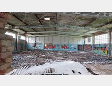 Verlassene Halle mit beschädigtem Holzfußboden und großflächiger Graffitikunst an den Wänden. Die Decke besteht aus Metallträgern, und große Fenster an den Seiten lassen Tageslicht herein.