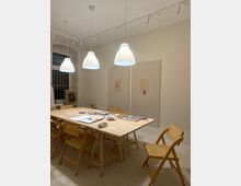 Heller Atelier- oder Seminarraum mit großem Holztisch und mehreren Holzstühlen, auf dem Tisch liegen Zeichen- und Bastelmaterialien. An der Decke hängen drei weiße Pendelleuchten und eine Schienenbeleuchtung, an der Wand sind große Pinnflächen und ein Fenster mit Gitter zu sehen.