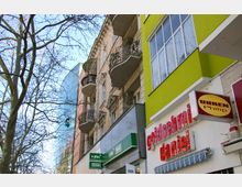 Mehrstöckige Gebäude entlang einer Straße, darunter ein altmodisches Gebäude mit verzierten Balkonen und ein moderneres Gebäude mit grüner Fassade. Im Erdgeschoss befinden sich Geschäfte, darunter ein Uhrenladen und ein Goldschmied, mit auffälligen Beschilderungen. Im Hintergrund ist eine Glasfassade zu sehen.