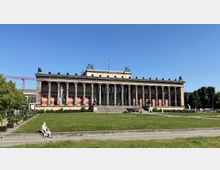Historisches Gebäude mit klassizistischer Architektur, bekannt als das Alte Museum in Berlin, mit einer Säulenfront und Statuen auf dem Dach. Im Vordergrund eine große Grünfläche mit Bäumen am Rand und mehreren Sitzgelegenheiten.