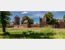 Blick über eine Wiese auf eine mittelalterliche Backsteinburg mit Ringmauer und mehreren Türmen, darunter ein hoher quadratischer Hauptturm. Im Hintergrund stehen große Laubbäume unter blauem Himmel, links ist ein Fachwerkgebäude an die Anlage angebaut.