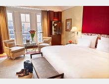 Elegantes Hotelzimmer mit einem großen Bett, rotem Kopfteil und weißen Kissen, einem Tisch mit zwei Sesseln vor bodentiefen Fenstern sowie einem Schrank mit Glasregalen. Auf dem Teppich stehen ein Paar braune Stiefel und eine dunkle Tasche.