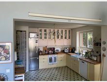 Helle Küche mit pastellgelben und rosa Schränken, Holzarbeitsplatten und einem Fliesenboden mit geometrischem Muster. Sichtbar sind ein großer Kühlschrank, ein Herd, Küchengeräte, ein Spülbecken unter einem Fenster sowie dekorative Elemente und eine hängende Lampe.