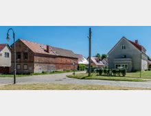 Straßenszene in einem Wohngebiet mit Kopfsteinpflaster und vereinzelten Häusern. Links ein altes Backsteingebäude mit Graffiti und rechts ein schlichtes Wohnhaus mit verputzter Fassade und eingezäuntem Garten, dazwischen ein Laternenmast und ein Strommast.