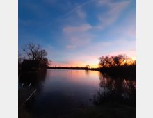 Seeufer bei Sonnenuntergang mit ruhigem Wasser und spiegelndem Himmel in Blau- und Orangetönen. Am linken Bildrand ist eine kleine Holzhütte auf Pfählen und ein Steg zu sehen, umgeben von kahlen Bäumen.