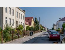 Straßenszene in einer Wohnsiedlung mit zweistöckigen Häusern, roten Ziegeldächern und Vorgärten, die teils mit Zäunen begrenzt sind; geparkte Autos säumen die asphaltierte Straße, und Strommasten verlaufen entlang der Gehwege.