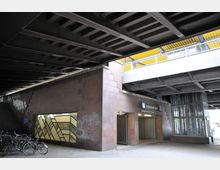 Eingang zur U-Bahn-Station "Bundesplatz" unter einer Brücke, mit brauner Steinverkleidung und einer verzierten Glaswand. Daneben befinden sich Fahrräder, Graffiti an der Wand und ein Aufzugsschacht mit Glasverkleidung ist sichtbar.